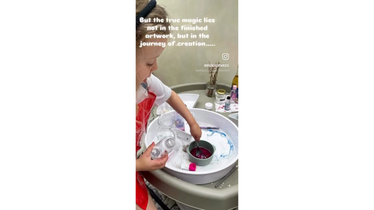 A group young girl seated at a table engaged in open-ended art: paint, brushes, and paper are spread out as each child explores freely with colours and textures.