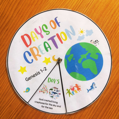 A circular educational spinner wheel titled 'Days of Creation' with colorful text, a blue and green Earth graphic, and a divided pointer pointing to the 'Day 5' section, placed on a wooden surface.