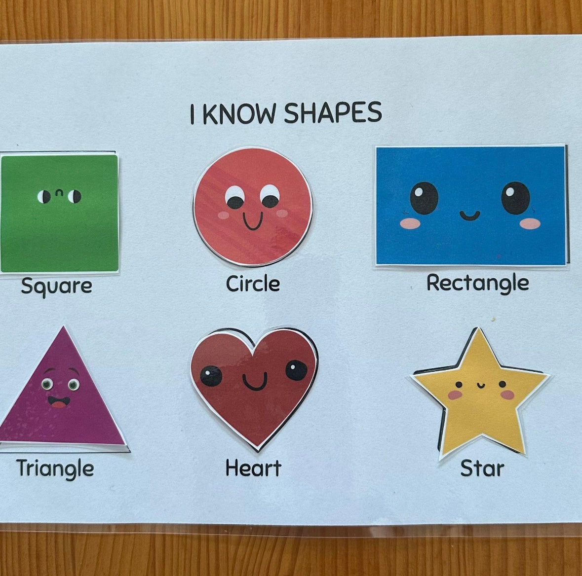 Educational card with shapes labeled 'Square', 'Circle', 'Rectangle', 'Triangle', 'Heart', and 'Star' on a wooden surface.