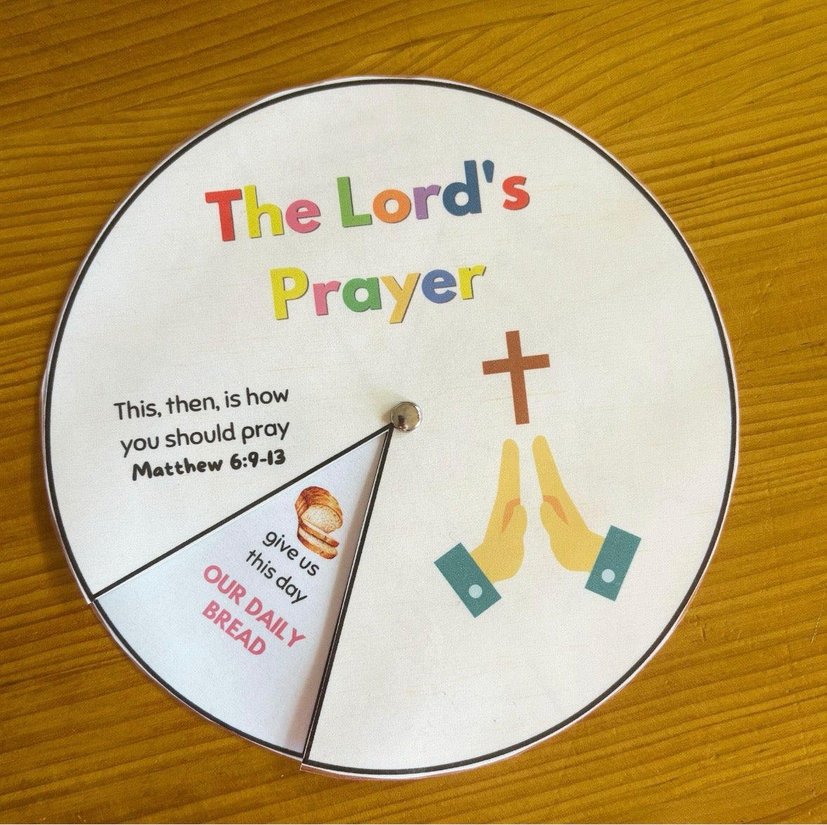 A circular printed prayer wheel or spinner labeled 'The Lord's Prayer' with a wooden cross, praying hands, and a small illustration of a loaf of bread, placed on a wooden tabletop.